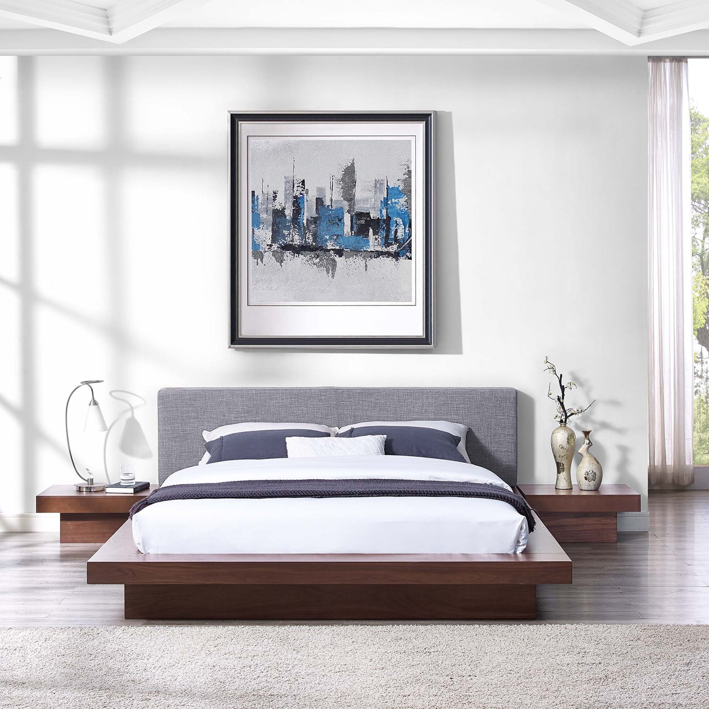 Walnut platform bed with gray upholstered headboard, white and navy bedding, and abstract blue cityscape art.