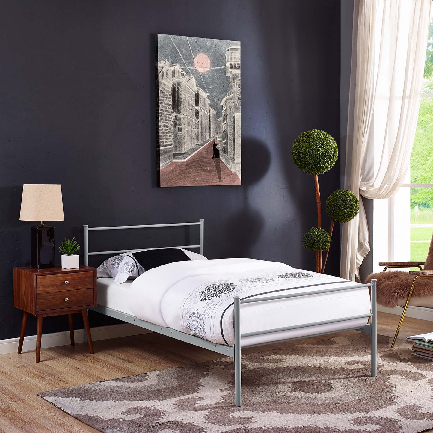 Gray metal twin bed frame with a white patterned duvet, on a dark wall, with a wooden nightstand, lamp, rug and greenery.