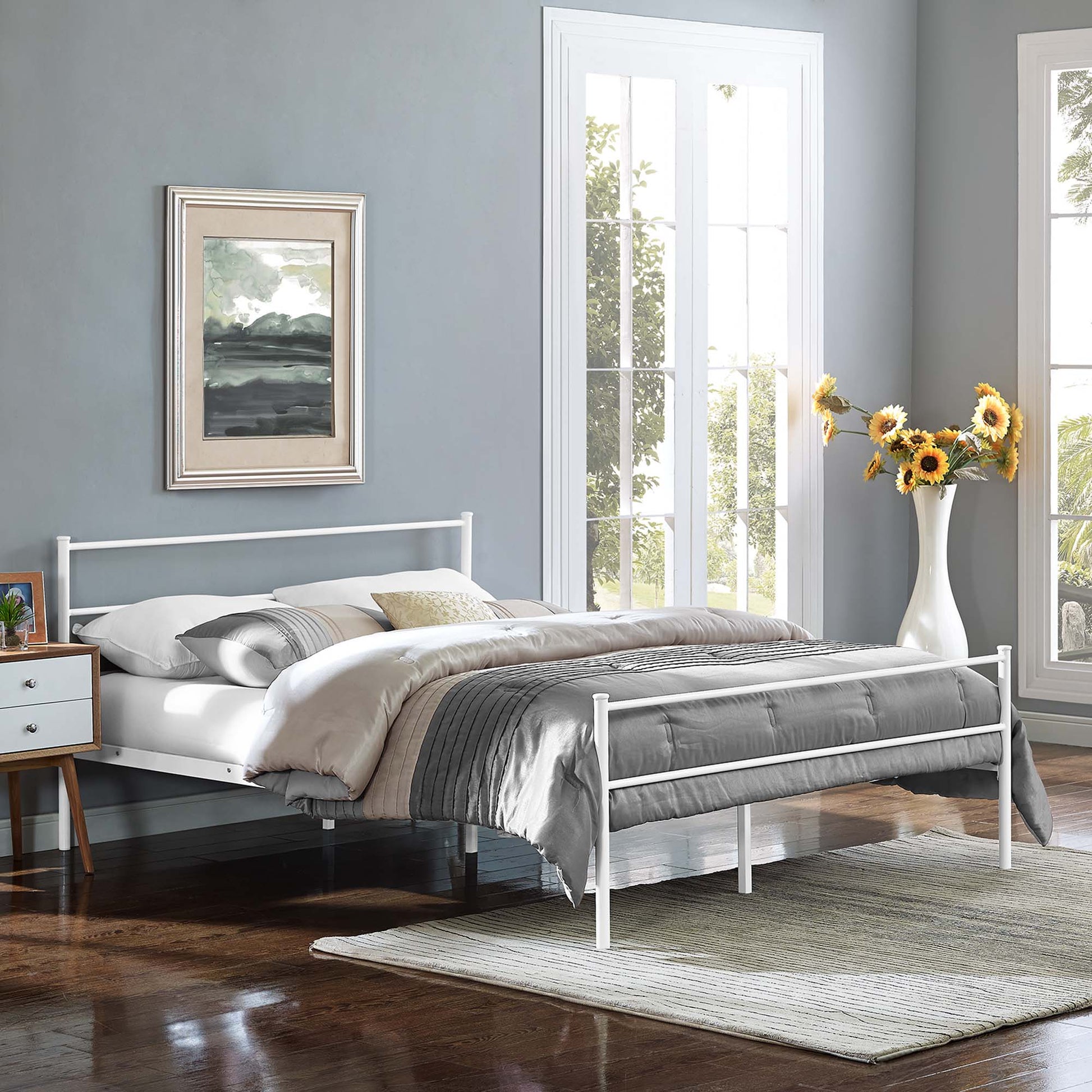 White metal bed frame with grey bedding in a light blue-gray bedroom, wood nightstand, sunflower vase, large windows.
