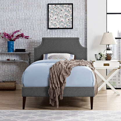 Gray fabric twin platform bed with squared tapered legs; light blue bedding and beige throw in a modern glam bedroom with a white nightstand.