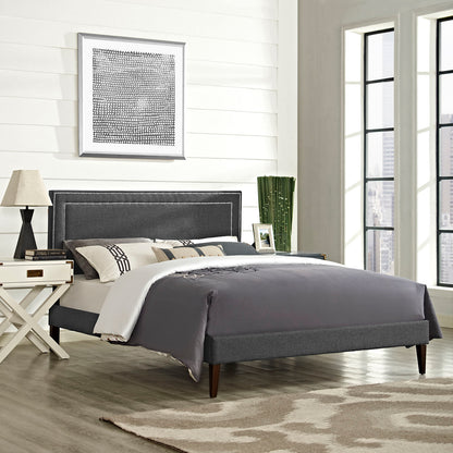 Grey upholstered full platform bed with squared tapered legs, charcoal comforter, white nightstand, beige lamp, and city-view windows.