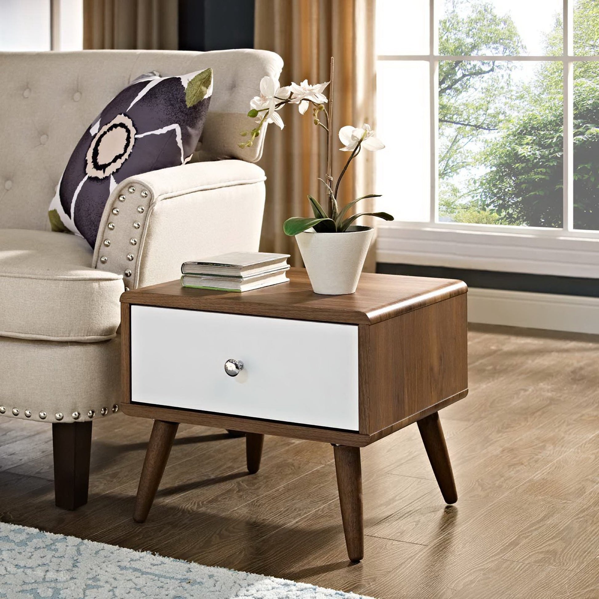 Walnut mid-century nightstand with a white front drawer, single knob, and tapered legs beside a beige sofa.