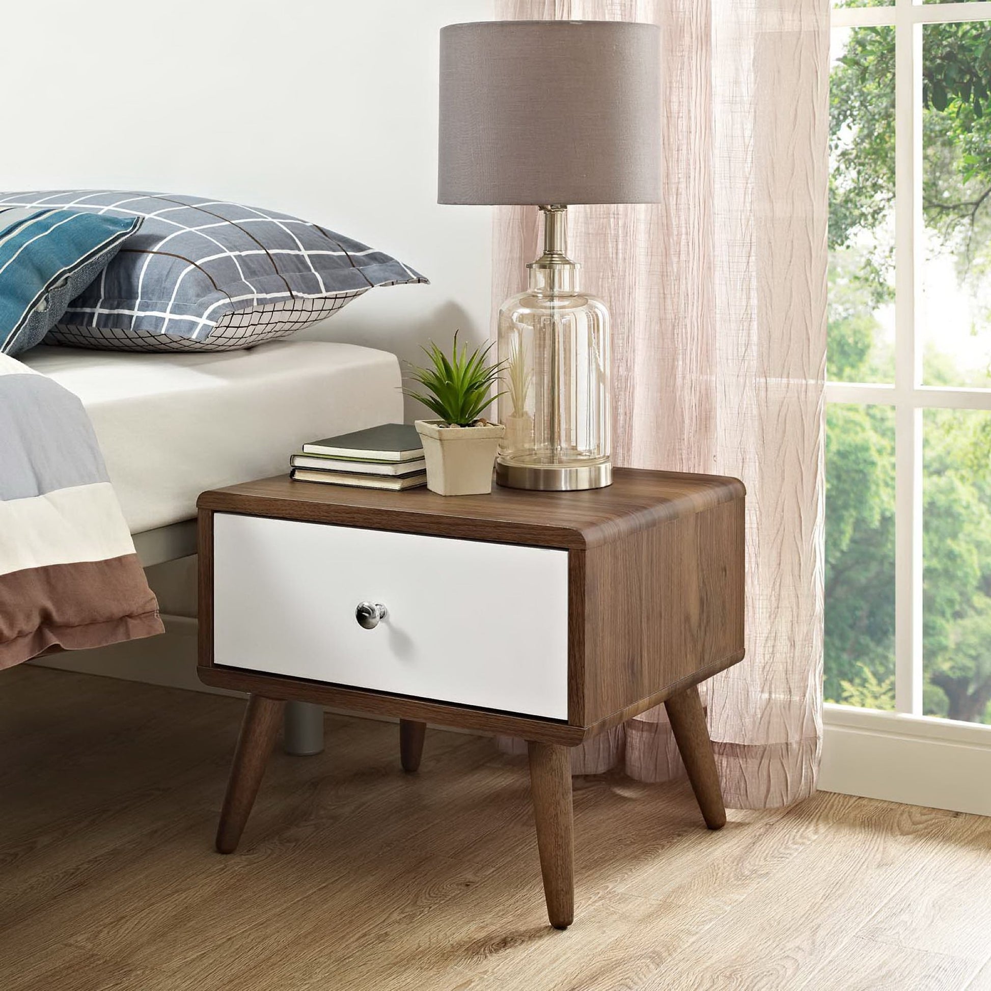 Walnut mid-century bedside nightstand with white front drawer, round knob, tapered legs; lamp, plant and books on top.