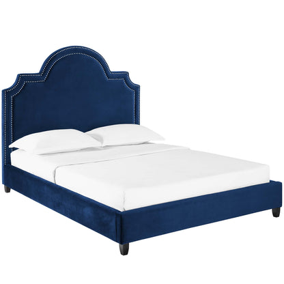 Navy blue velvet upholstered bed with a curved, studded headboard and low frame, white bedding.