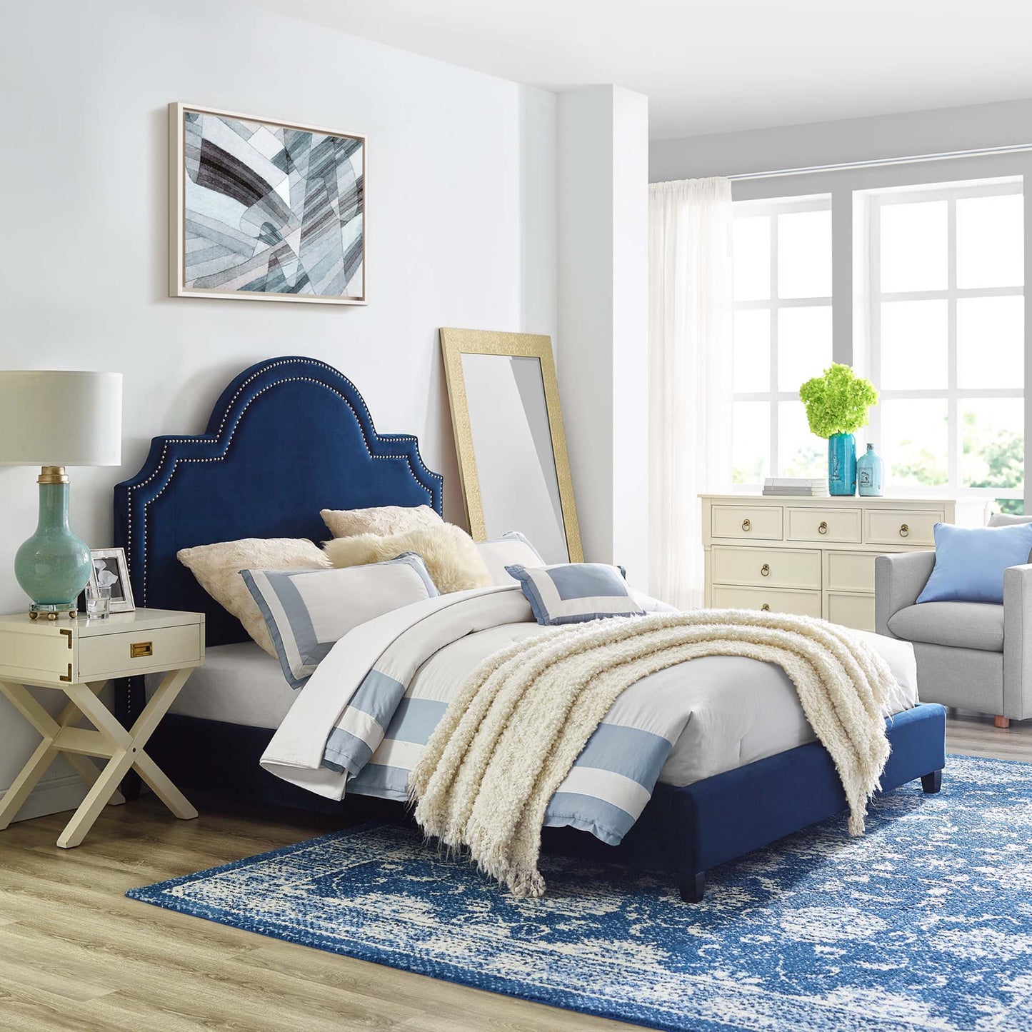 Navy upholstered bed with curved, studded headboard, white blue striped bedding, cream throw in a bright coastal bedroom.