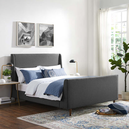 Gray fabric upholstered bed with a tall headboard, blue-and-white bedding, gold-accent lamps, plant, and abstract wall art.