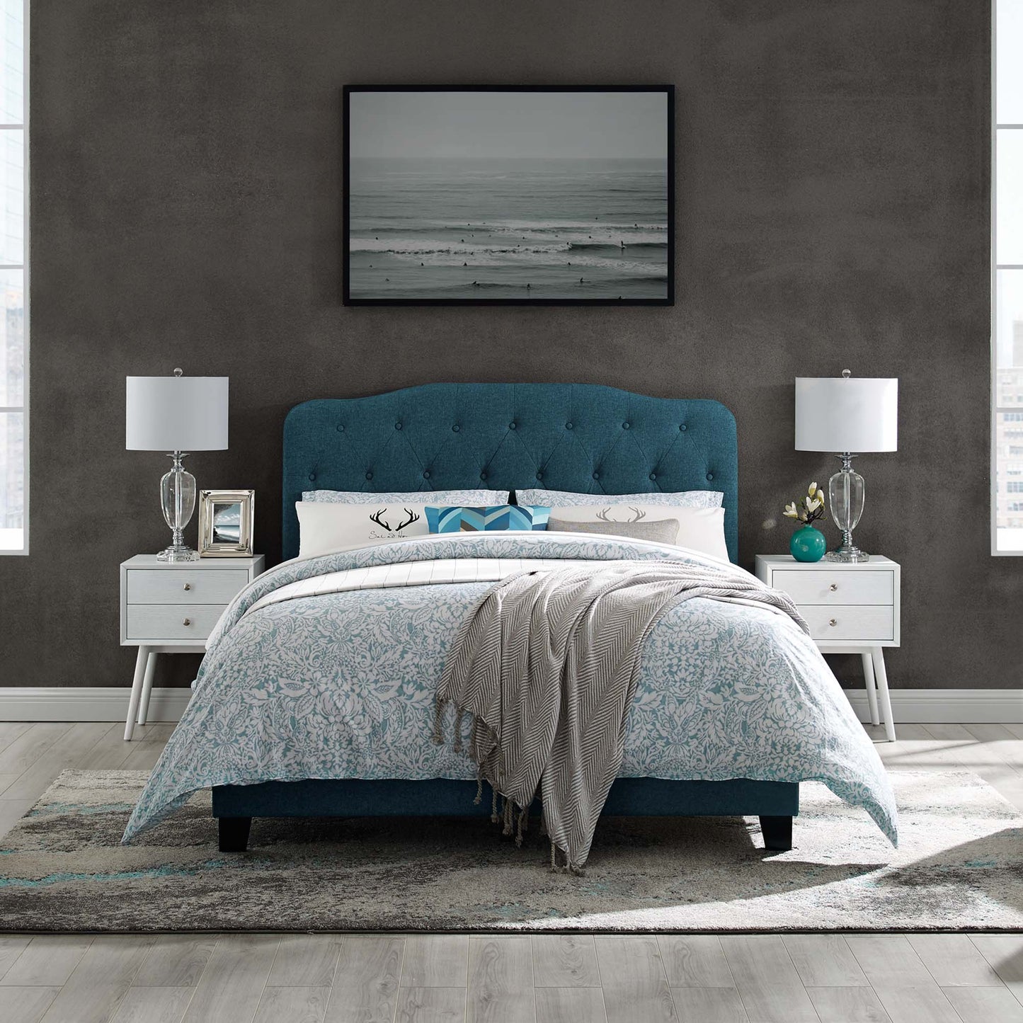 Teal blue tufted upholstered bed with white nightstands and glass lamps; ocean-photo art, blue floral duvet, gray throw.