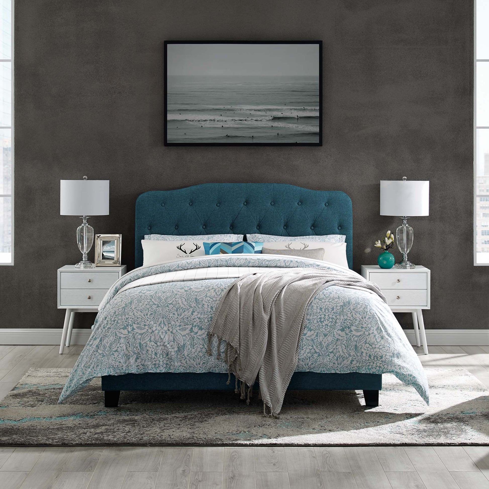 Teal blue tufted upholstered bed with white nightstands and glass lamps; ocean-photo art, blue floral duvet, gray throw.