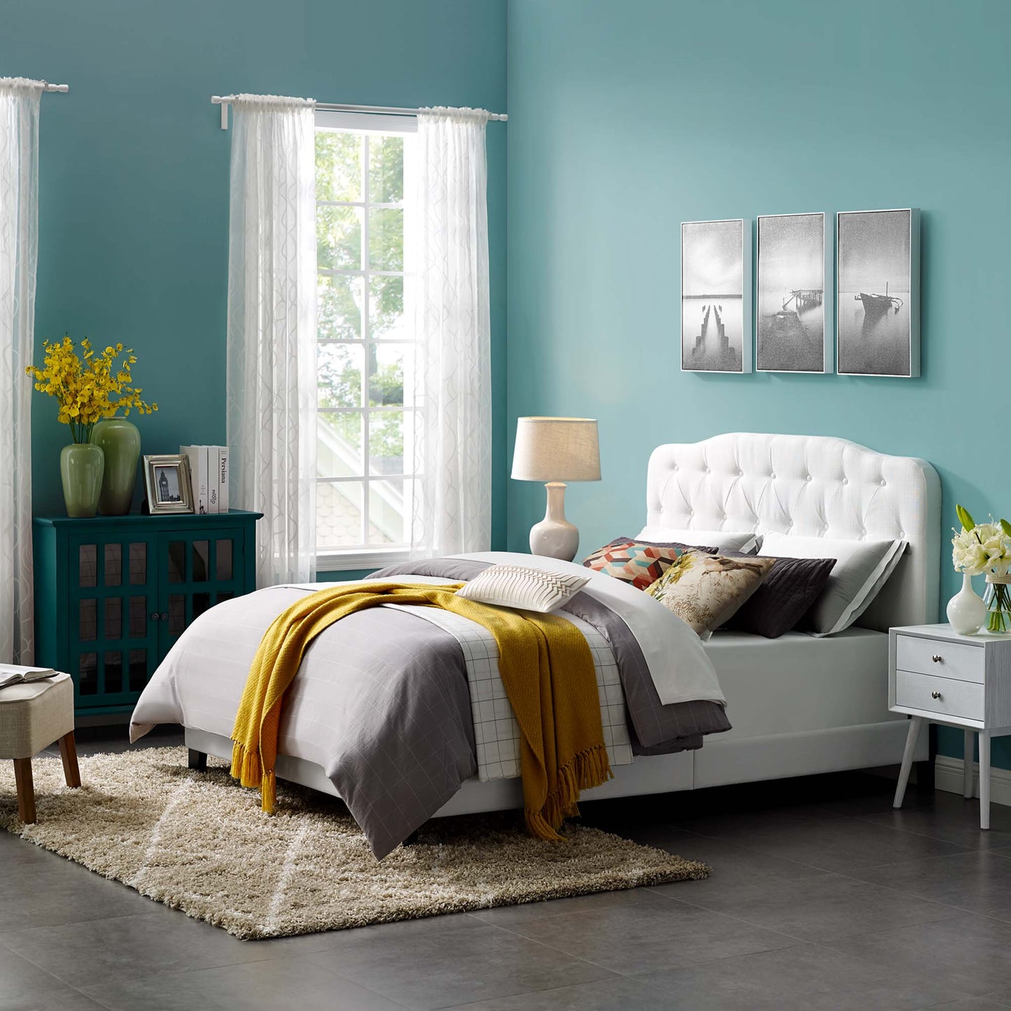 Teal-walled bedroom with white tufted bed, gray bedding, mustard throw, white nightstand, turquoise cabinet, triptych art, cream rug.