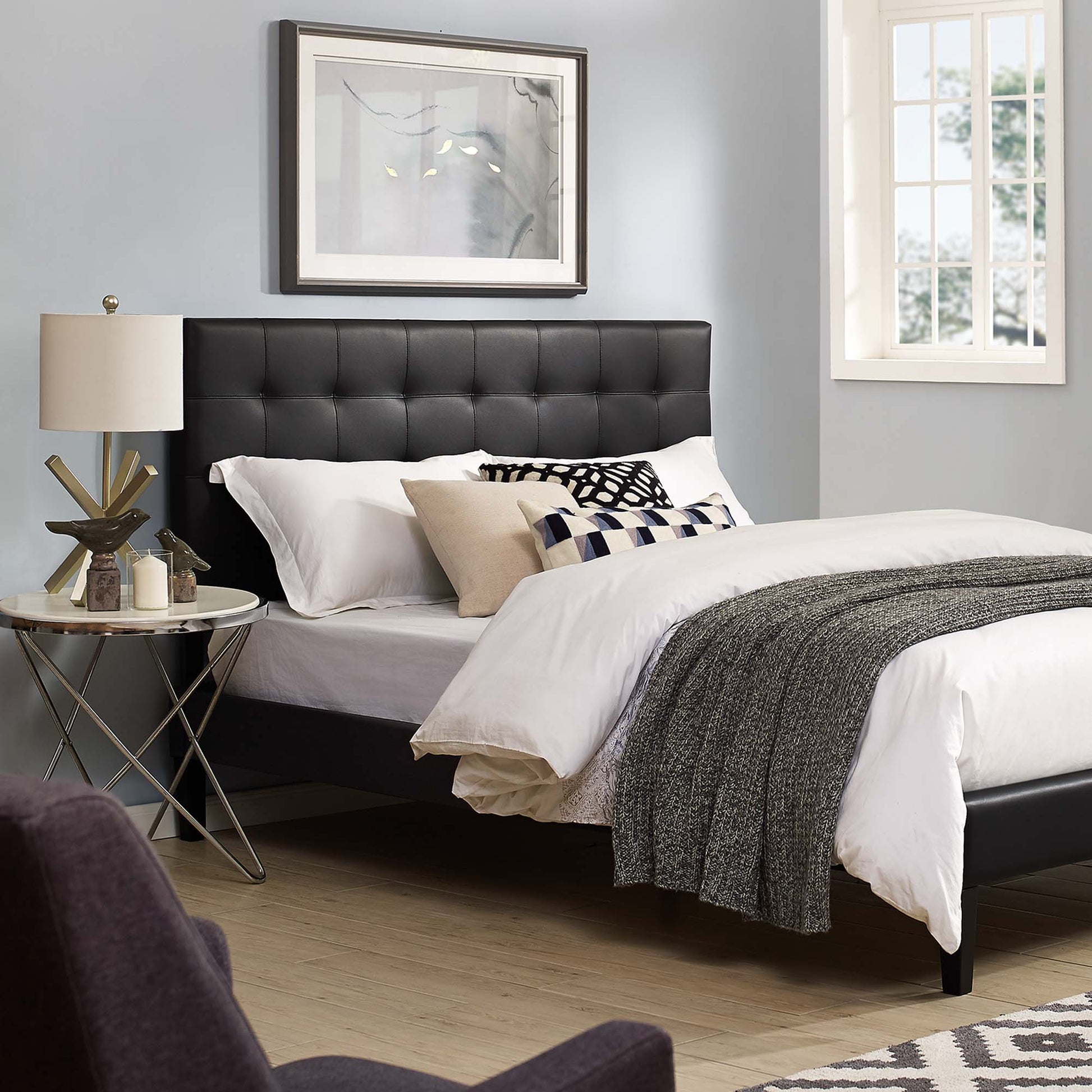 Black tufted headboard with white linens and gray throw; beige pillows, round glass side table, lamp, framed art, light blue walls.