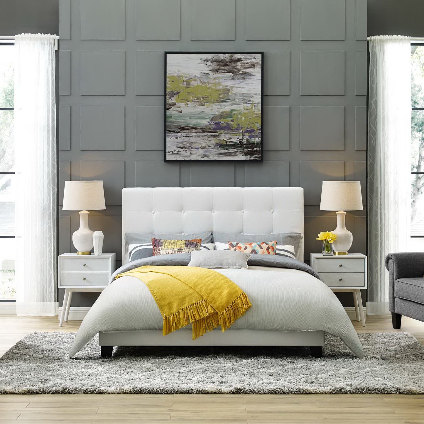 White tufted fabric queen platform bed with gray paneled wall, yellow throw blanket, matching nightstands & lamps, abstract wall art.