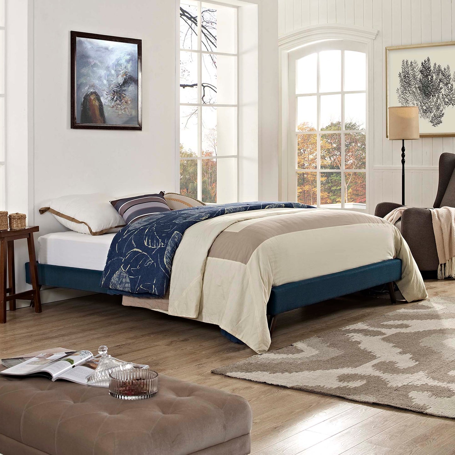 Teal upholstered bed frame with beige duvet and navy quilt, beige tufted ottoman, light wood floor in a bright coastal bedroom.