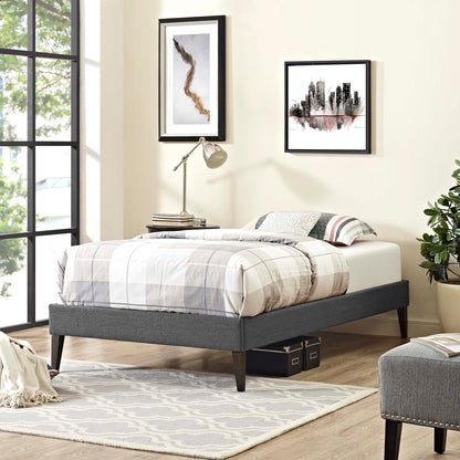 Gray upholstered platform bed with plaid bedding, squared tapered legs, modern bedroom with framed art, lamp, rug, and plant