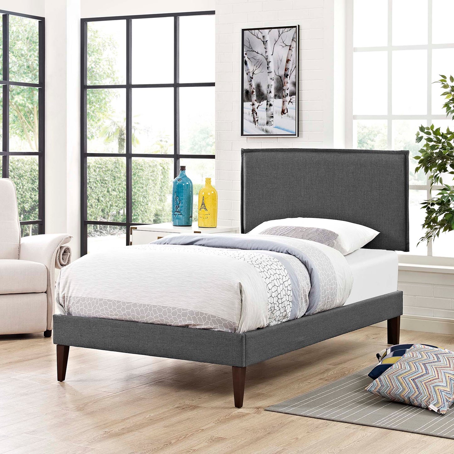 Grey upholstered platform bed with tall headboard and tapered dark wood legs in a bright modern bedroom.
