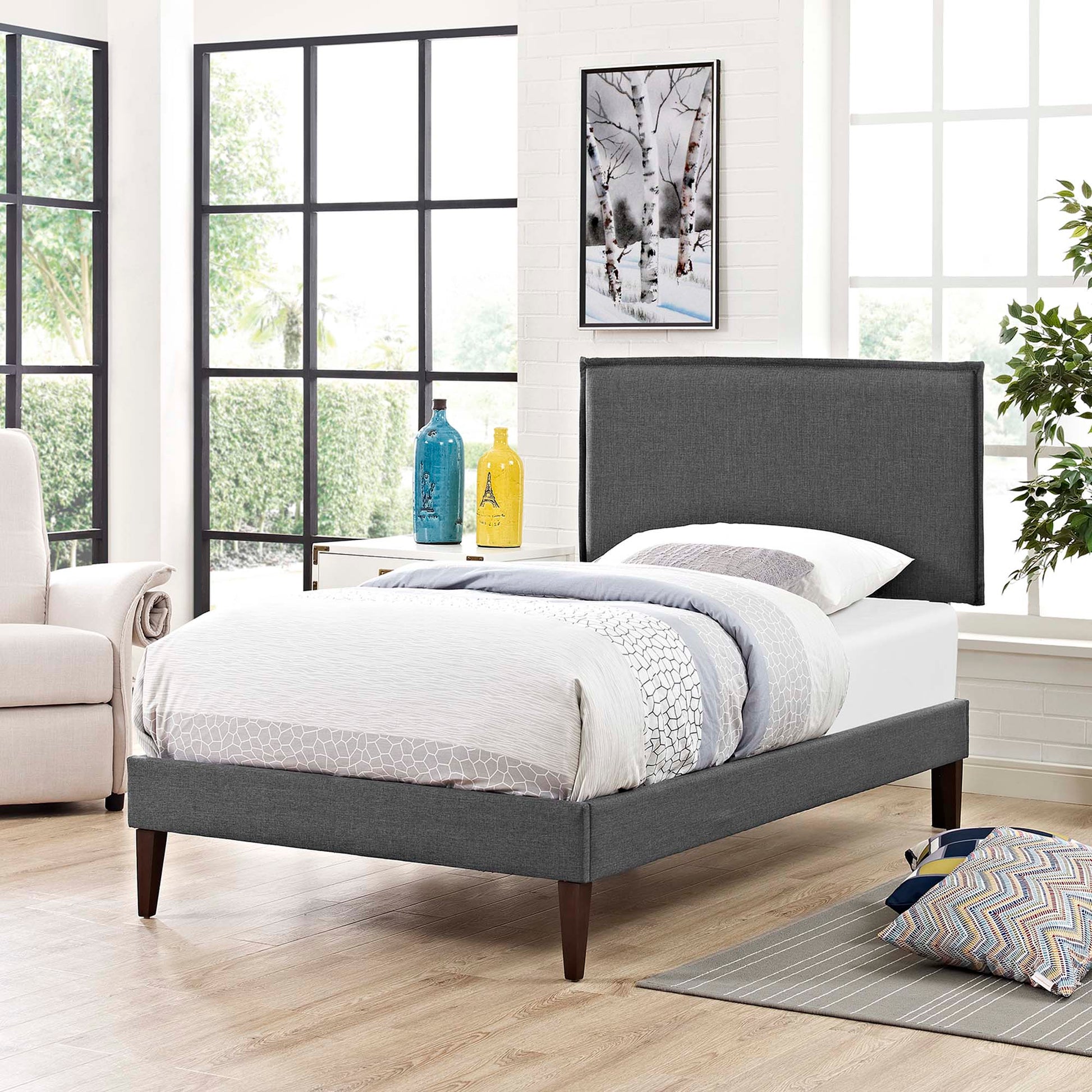 Grey upholstered platform bed with tall headboard and tapered dark wood legs in a bright modern bedroom.