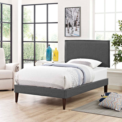 Grey upholstered platform bed with tall headboard and tapered dark wood legs in a bright modern bedroom.