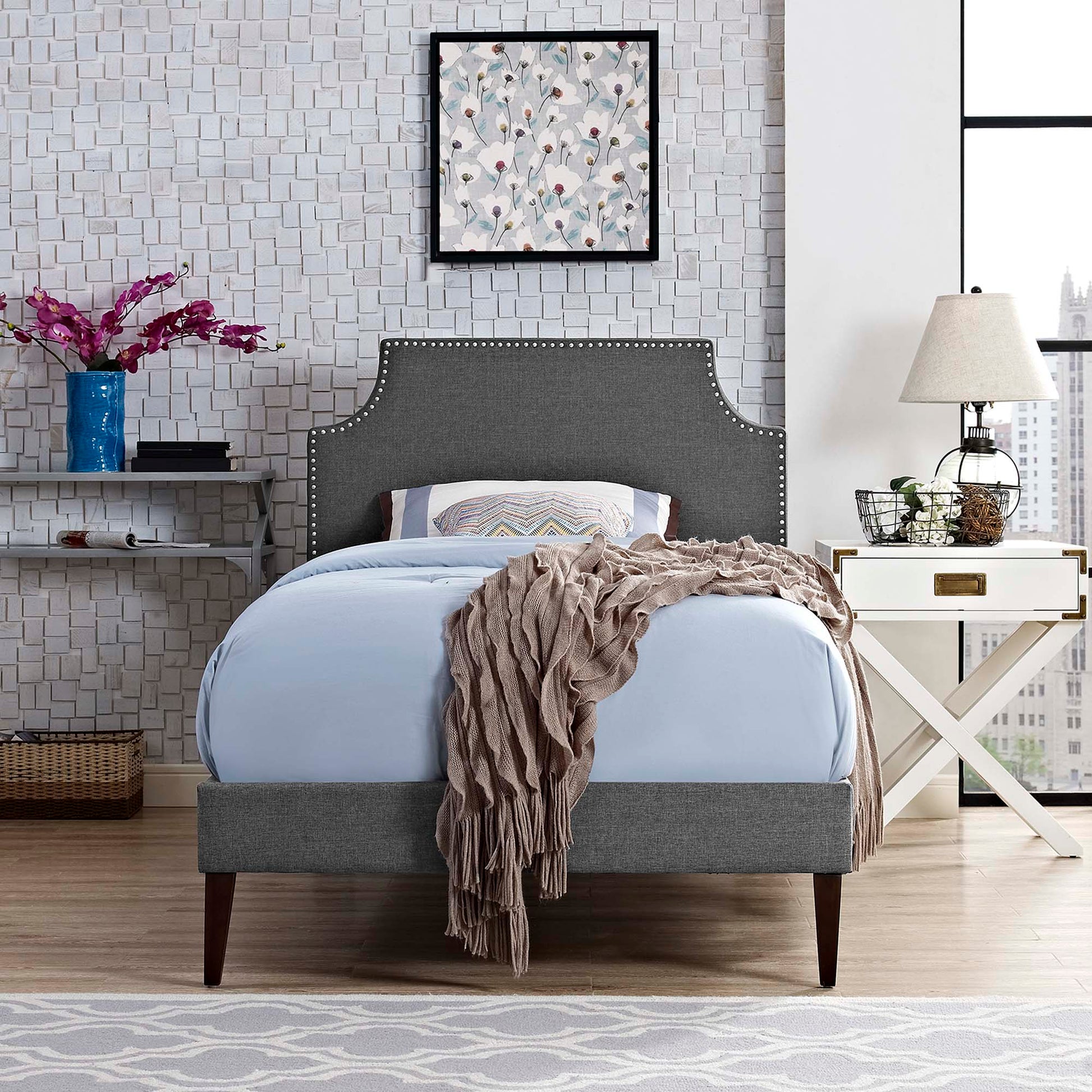 Grey upholstered bed with nailhead trim, curved headboard, light blue bedding, taupe throw, white nightstand, modern bedroom.