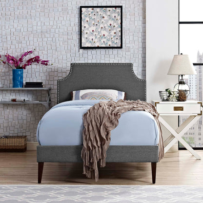 Grey upholstered bed with nailhead trim, curved headboard, light blue bedding, taupe throw, white nightstand, modern bedroom.
