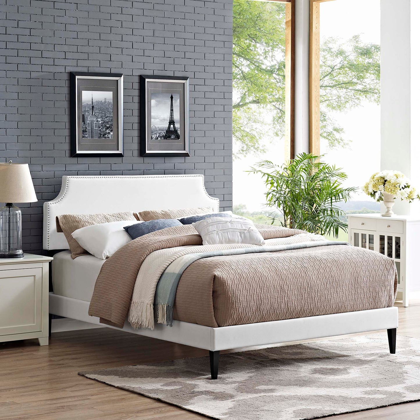 White vinyl platform bed with curved headboard, beige quilt, blue throw, gray brick wall, two black-framed city photos.