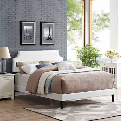 White vinyl platform bed with curved headboard, beige quilt, blue throw, gray brick wall, two black-framed city photos.