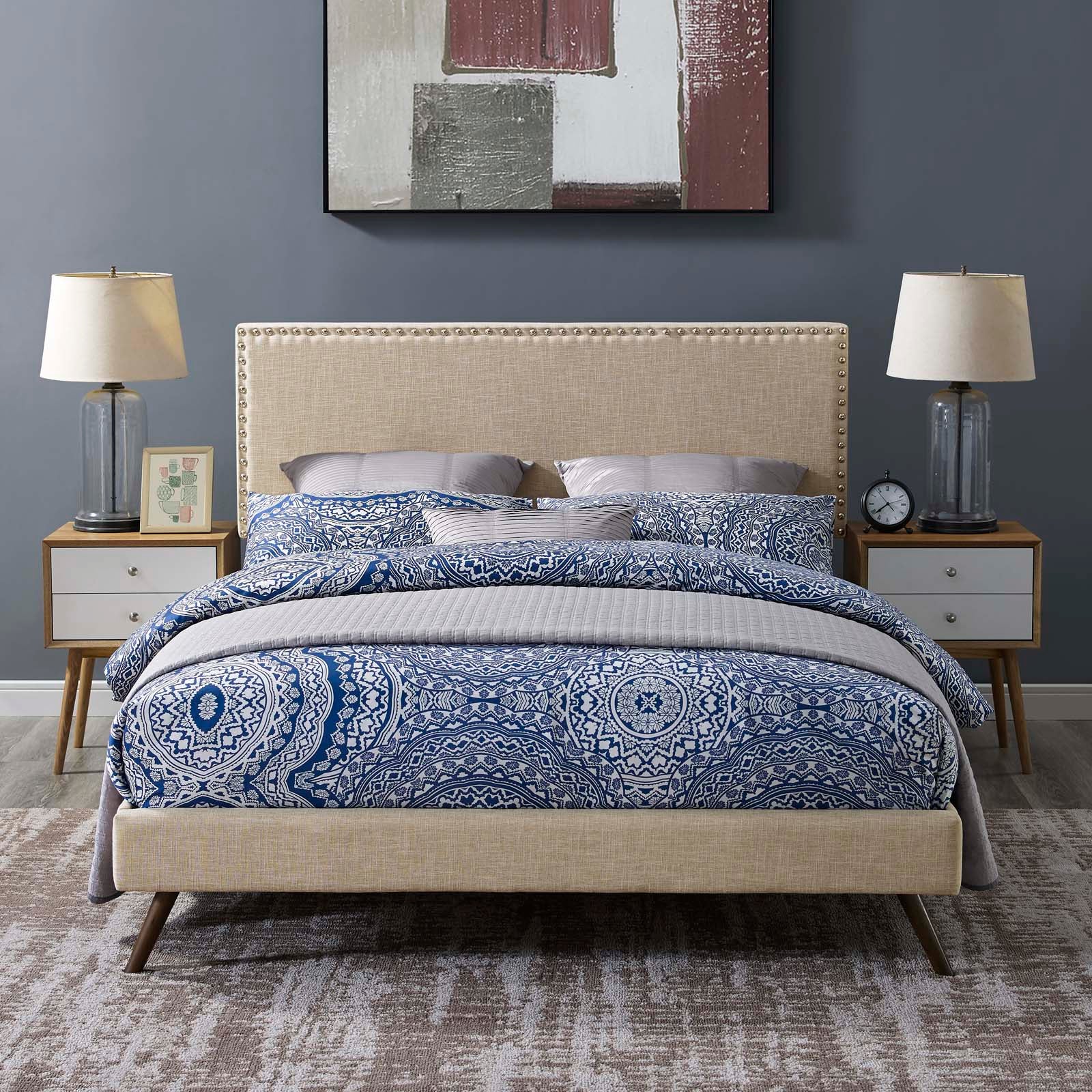 Beige fabric queen platform bed with round splayed legs, blue patterned bedding, white nightstands, glass lamps, blue-gray walls.