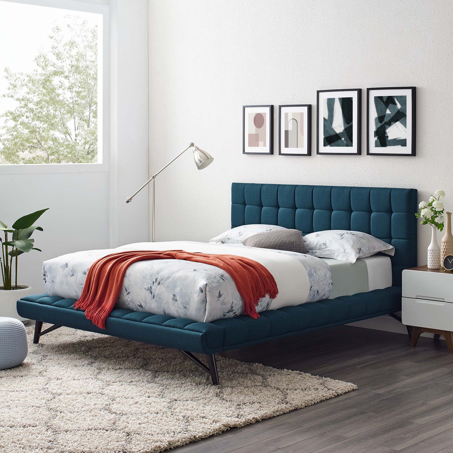 Blue upholstered queen platform bed with a tufted headboard and footboard, floral bedding and an orange throw in a modern bedroom.