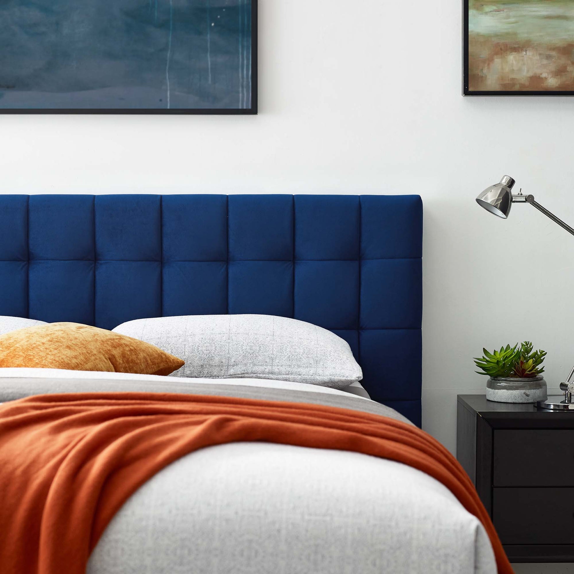 Navy tufted platform bed with white linens and a burnt orange throw; dark nightstand, silver lamp, and a succulent.