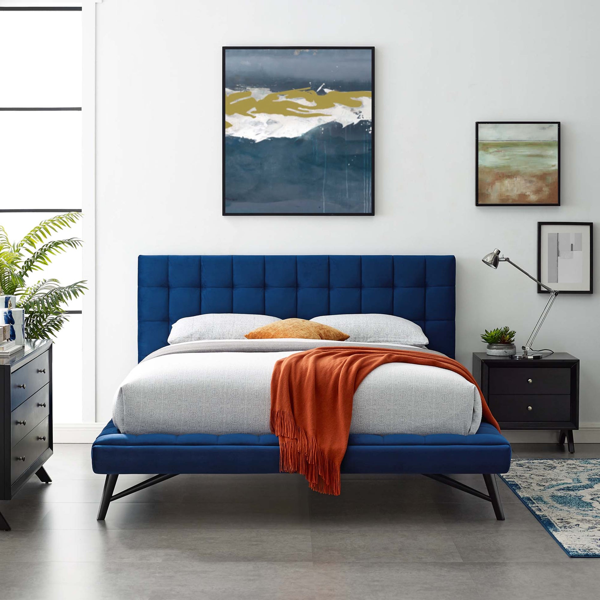 Navy tufted platform bed with grey bedding and orange throw in a modern room with black nightstands and a blue rug.