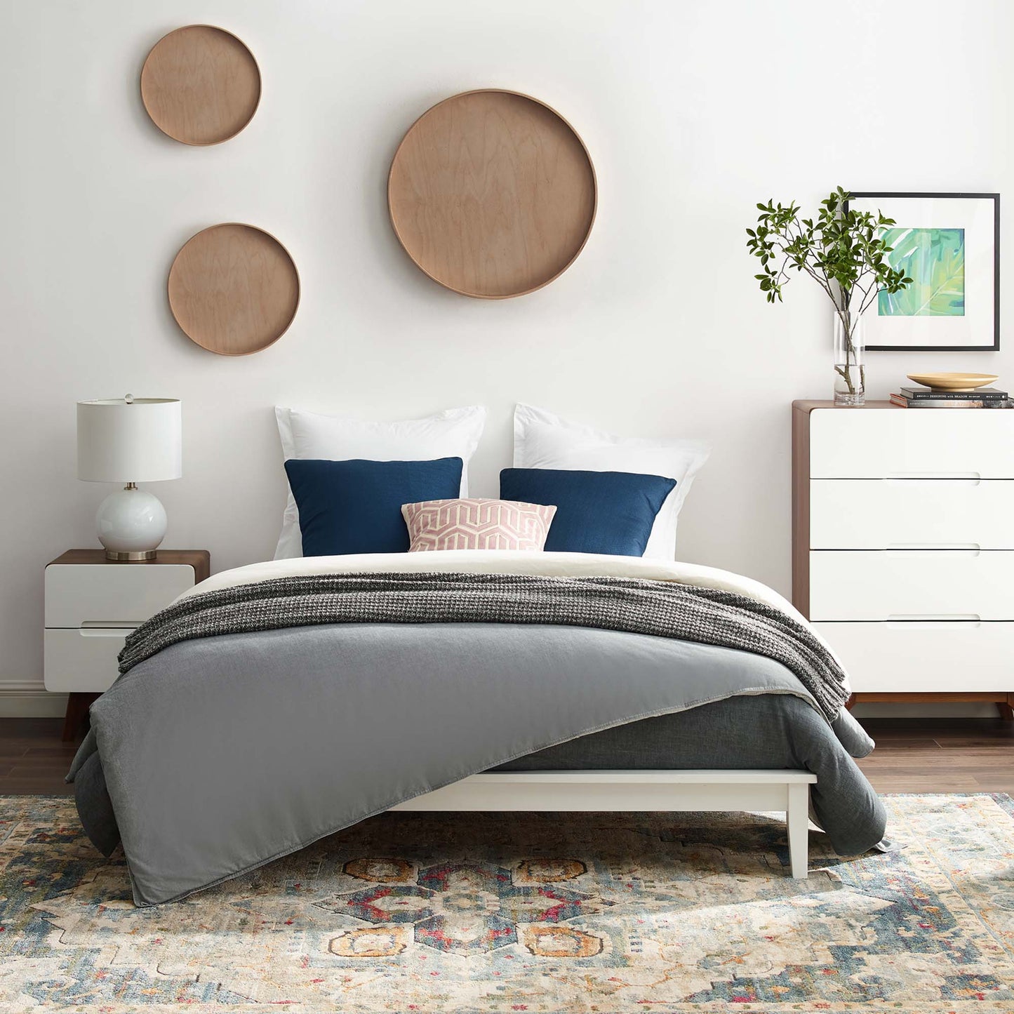 Modern bedroom featuring a white full-size platform bed frame, a gray duvet, white & navy pillows, circular wood wall decor, white dressers, and a rug.