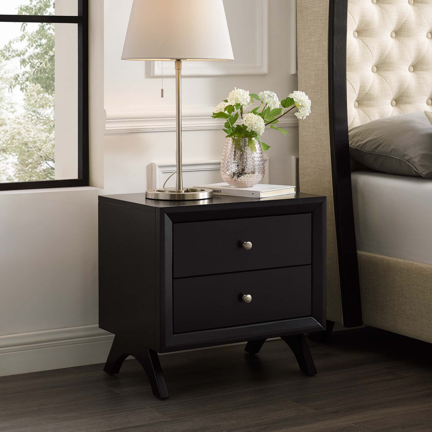 Cappuccino two-drawer nightstand with silver knobs and tapered legs, lamp on top with a vase of white flowers beside a tufted bed.