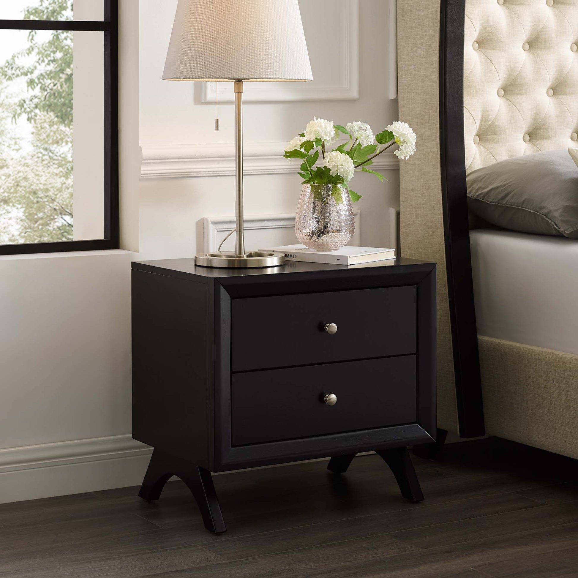 Cappuccino two-drawer nightstand with silver knobs and tapered legs, lamp on top with a vase of white flowers beside a tufted bed.