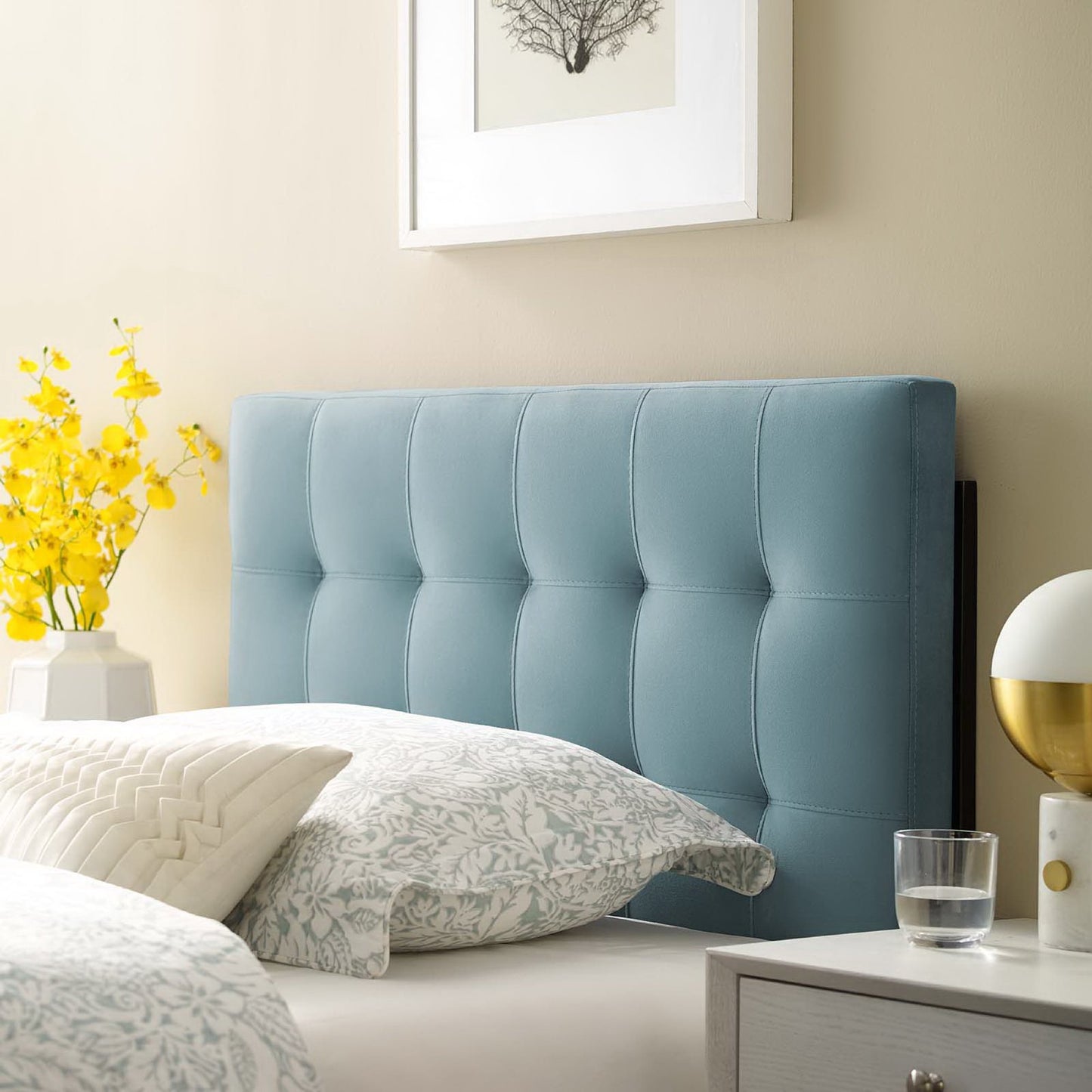 Blue tufted upholstered headboard in a beige bedroom with patterned gray bedding, yellow flowers, and framed art.