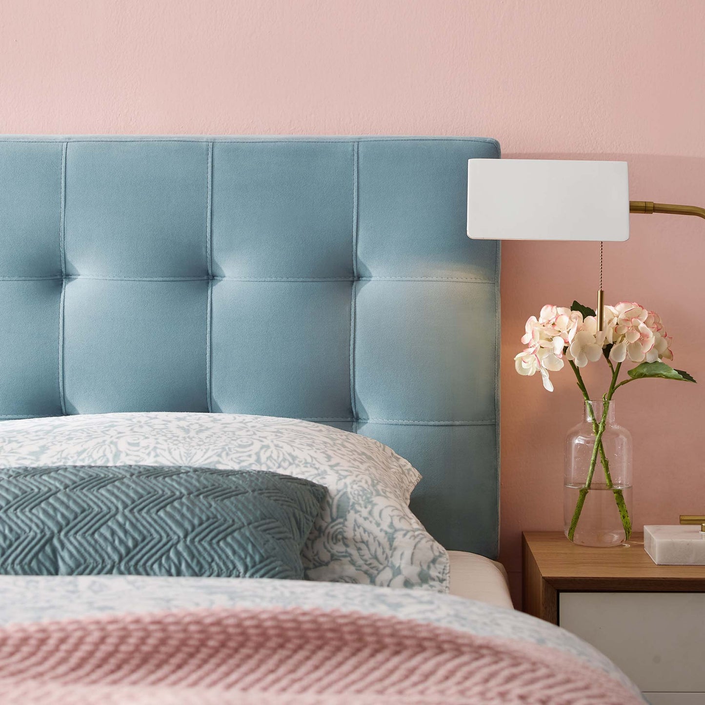 Blue tufted headboard on pink wall, nightstand with white lamp and glass vase of white orchids, patterned bedding.