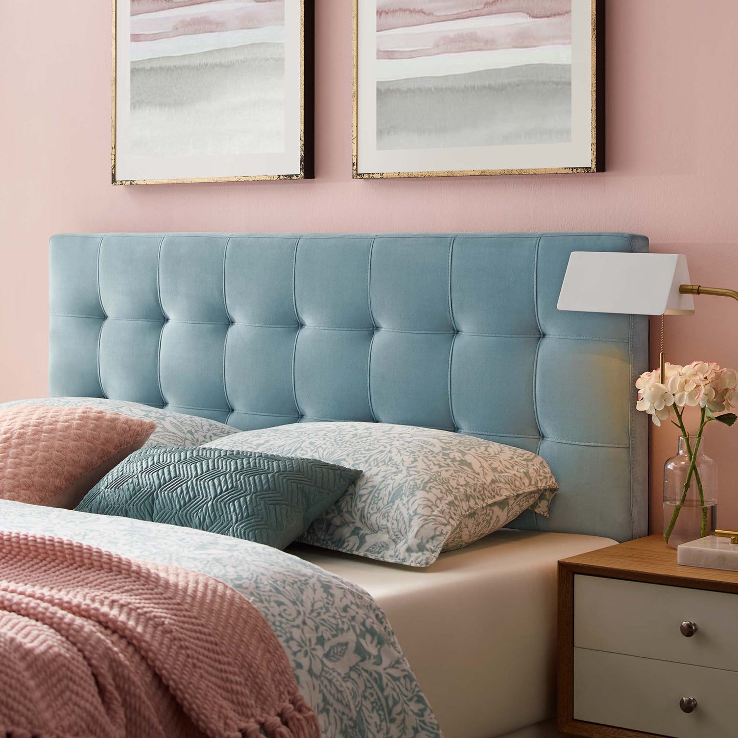 Blue tufted upholstered headboard on pink wall with pastel bedding, pink throw, pillows, wooden nightstand and lamp.
