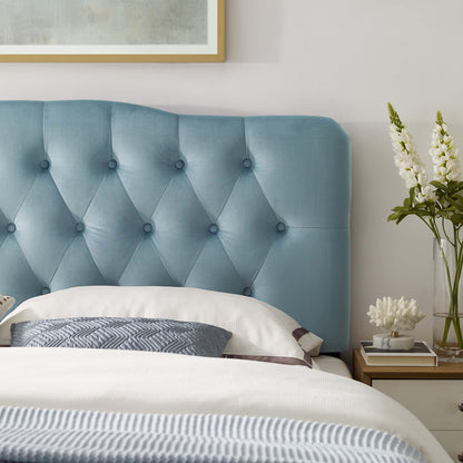 Blue tufted headboard bed with white linens, grey patterned pillow, wooden nightstand, and vase of white flowers.