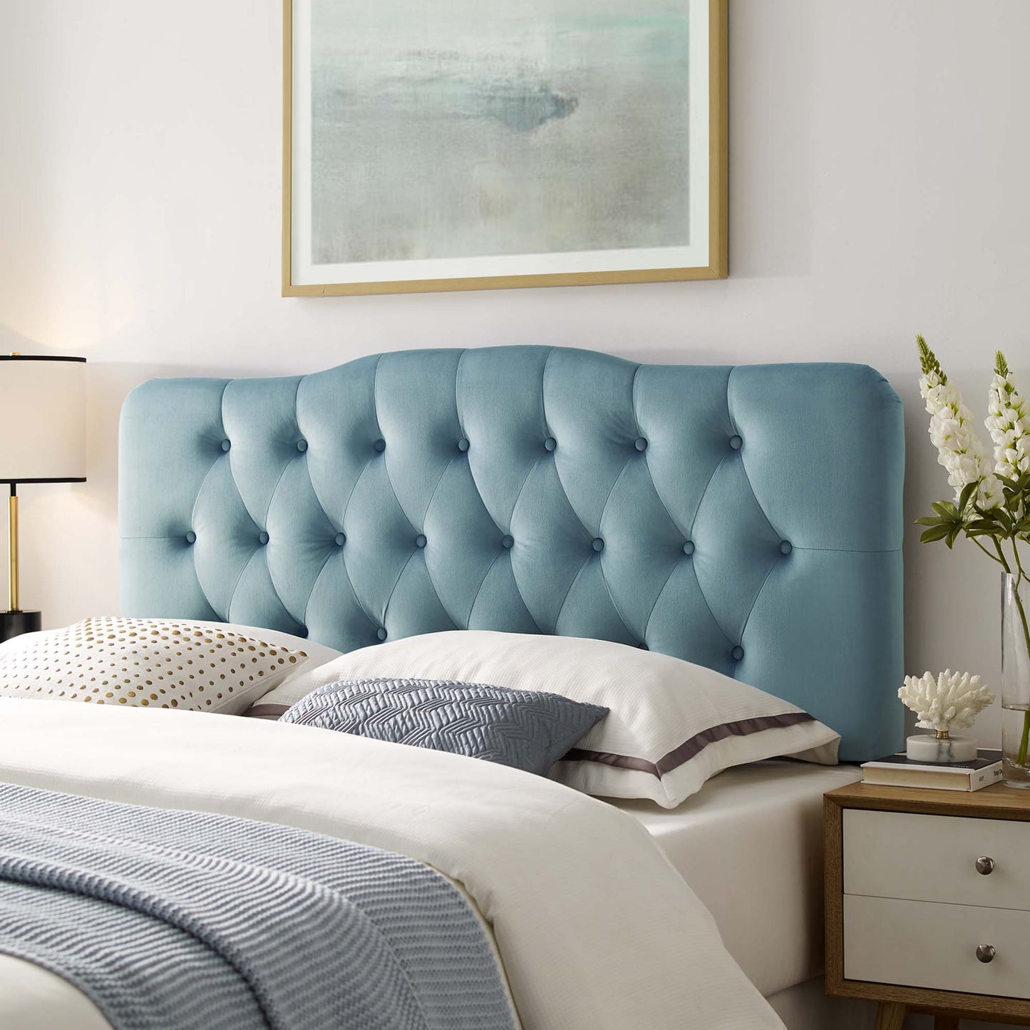 Modern coastal bedroom with a light blue tufted velvet headboard, white bedding, accent pillows, and a nightstand with lamp.