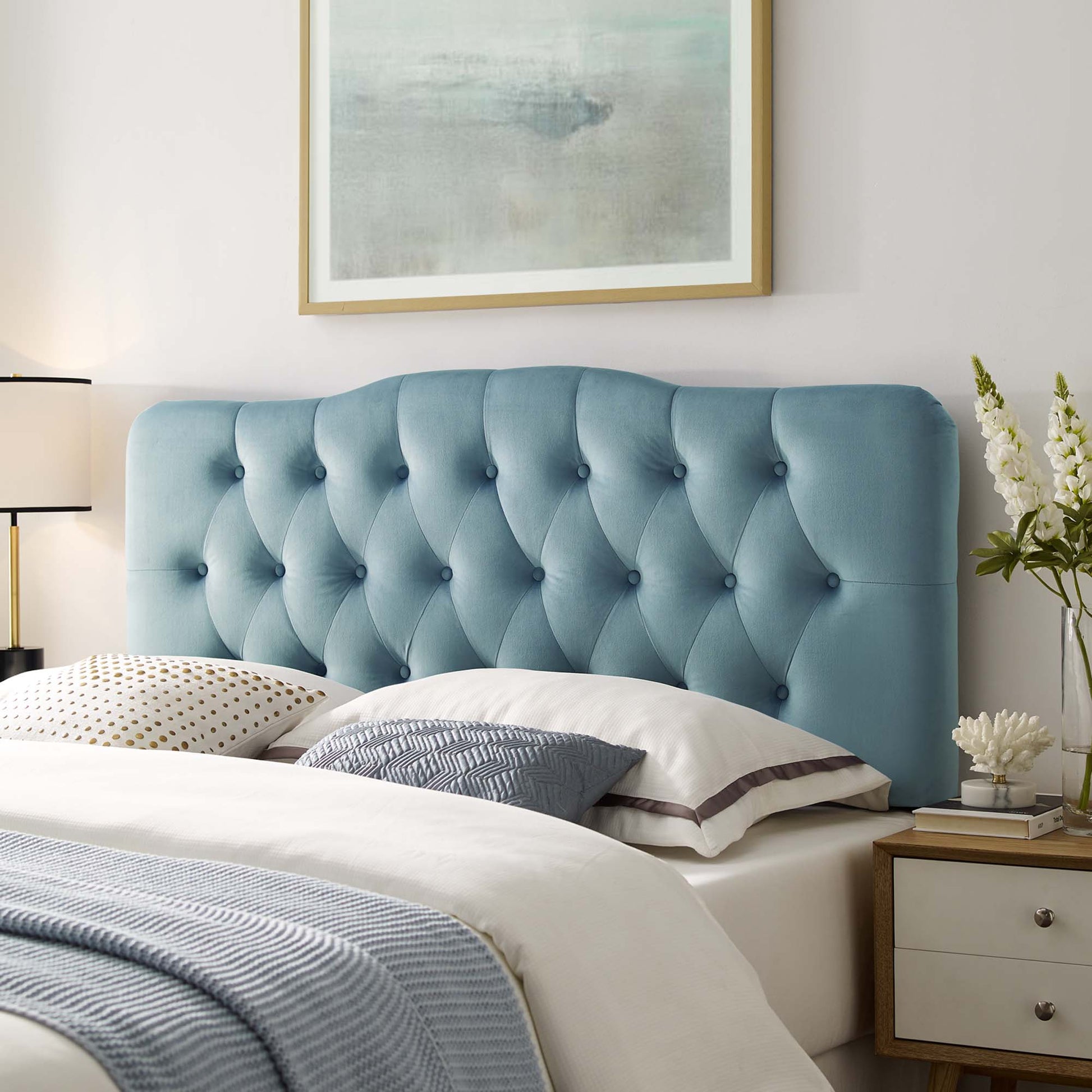 Modern coastal bedroom with a light blue tufted velvet headboard, white bedding, accent pillows, and a nightstand with lamp.