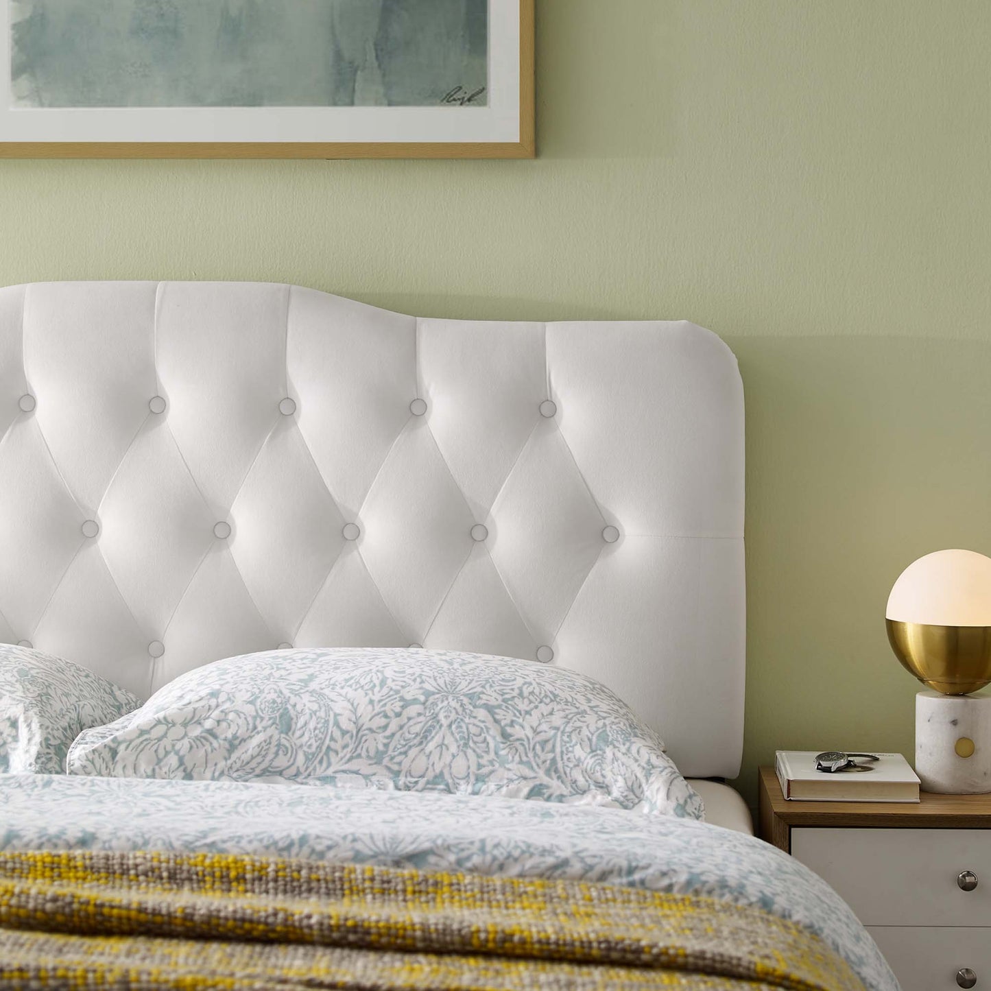 White tufted upholstered headboard with button detailing, light blue patterned bedding, pale green wall, nightstand with gold lamp.