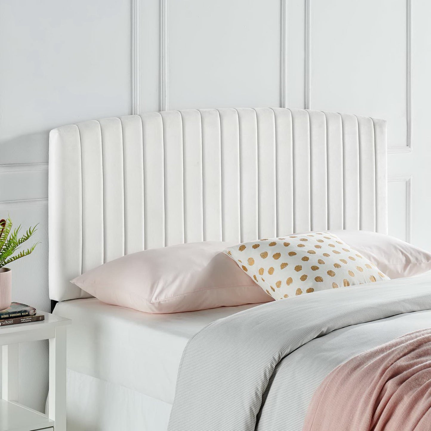 White upholstered headboard with vertical channels, pink pillows, pale gray blanket, white nightstand with plant.