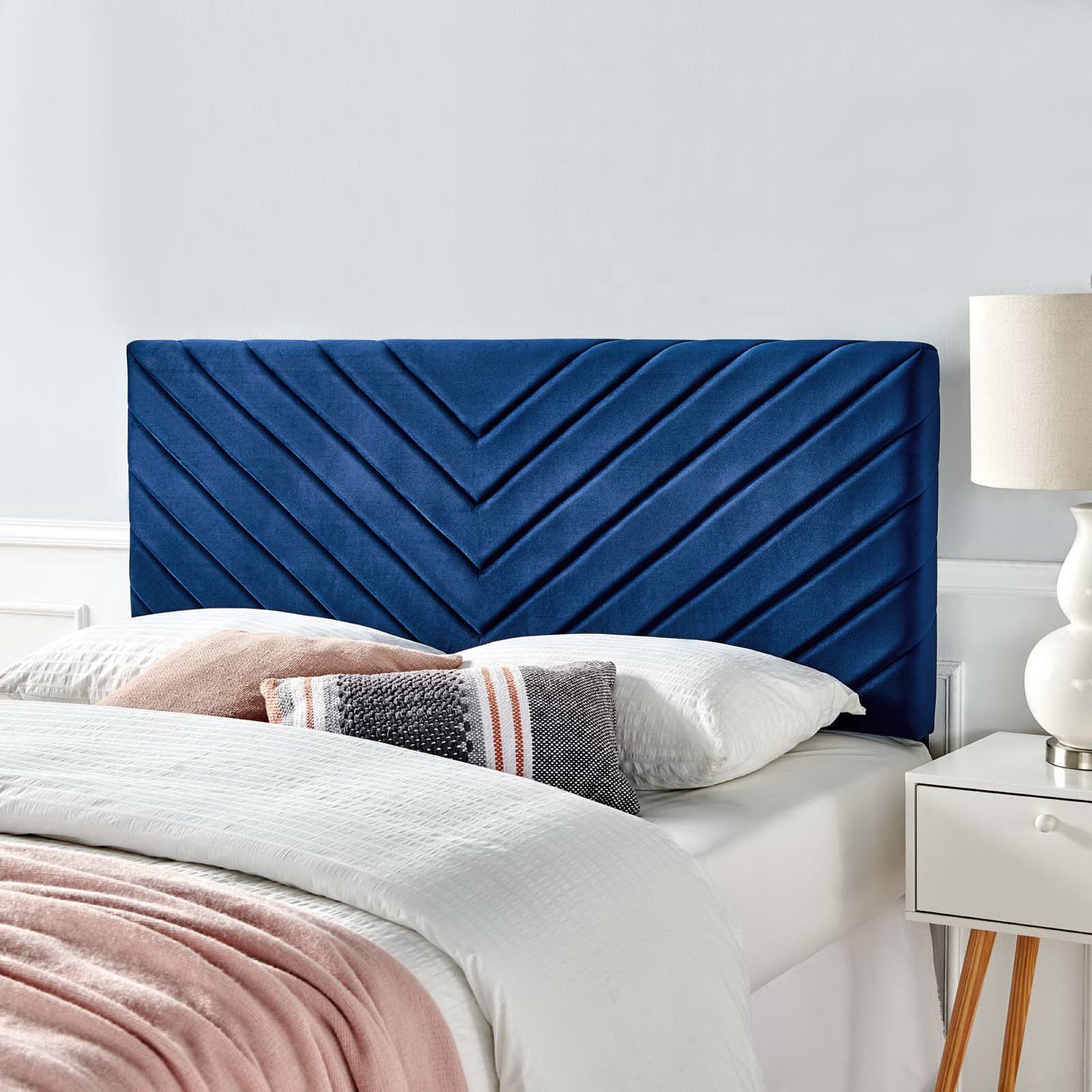 Navy blue velvet angular channel tufted headboard on white bed with white linens, pink throw, gray striped pillow, bedside lamp.