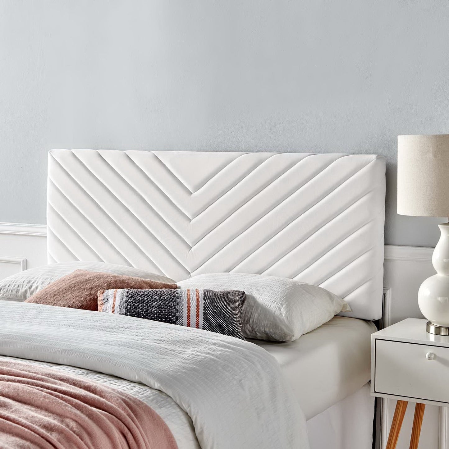 White upholstered headboard with angular channel tufting, shown with bed linens, pink throw, striped pillow, & white nightstand with a beige lamp.