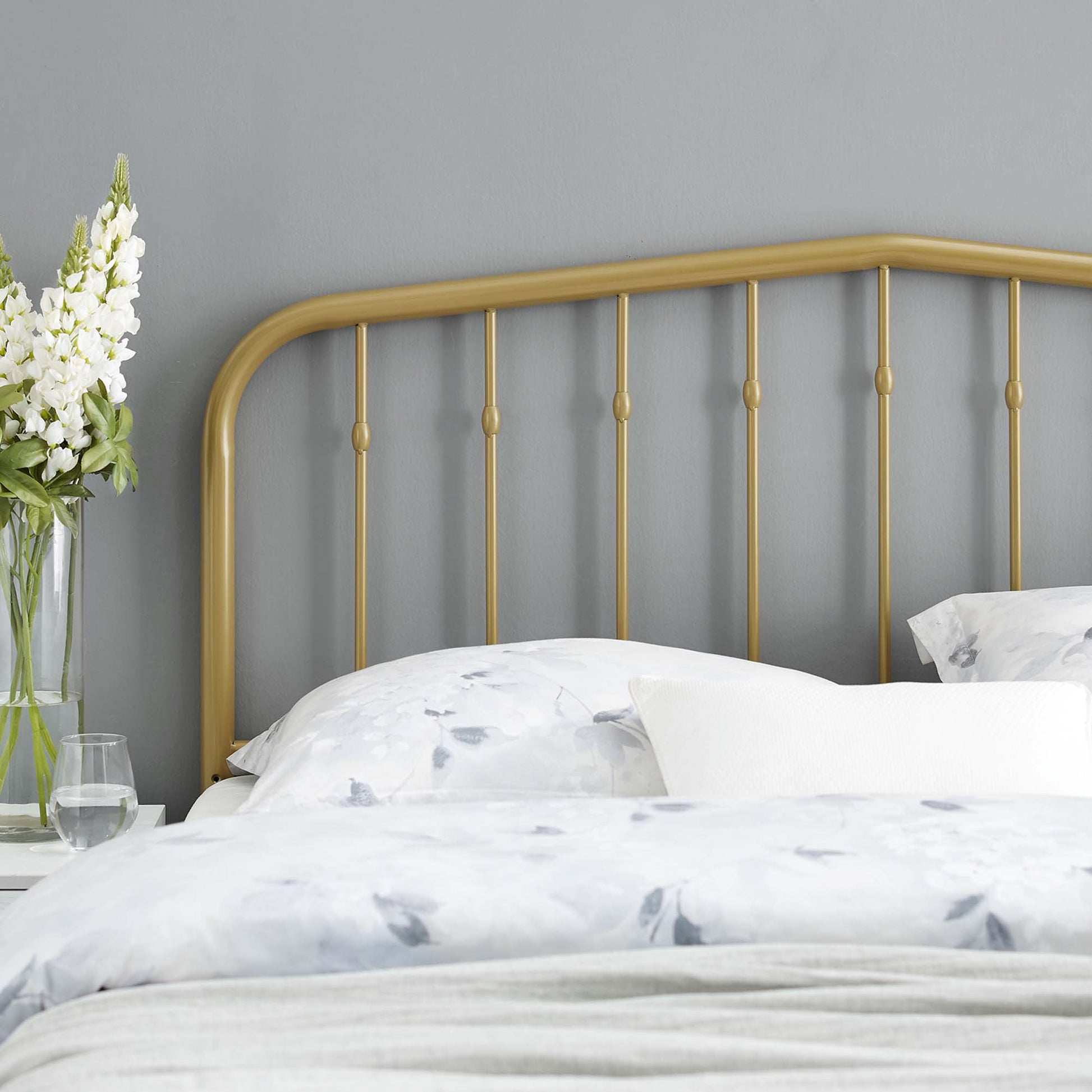 Yellow metal headboard with a curved top rail and vertical posts, white floral bedding, gray wall, and a vase of white flowers on the nightstand.