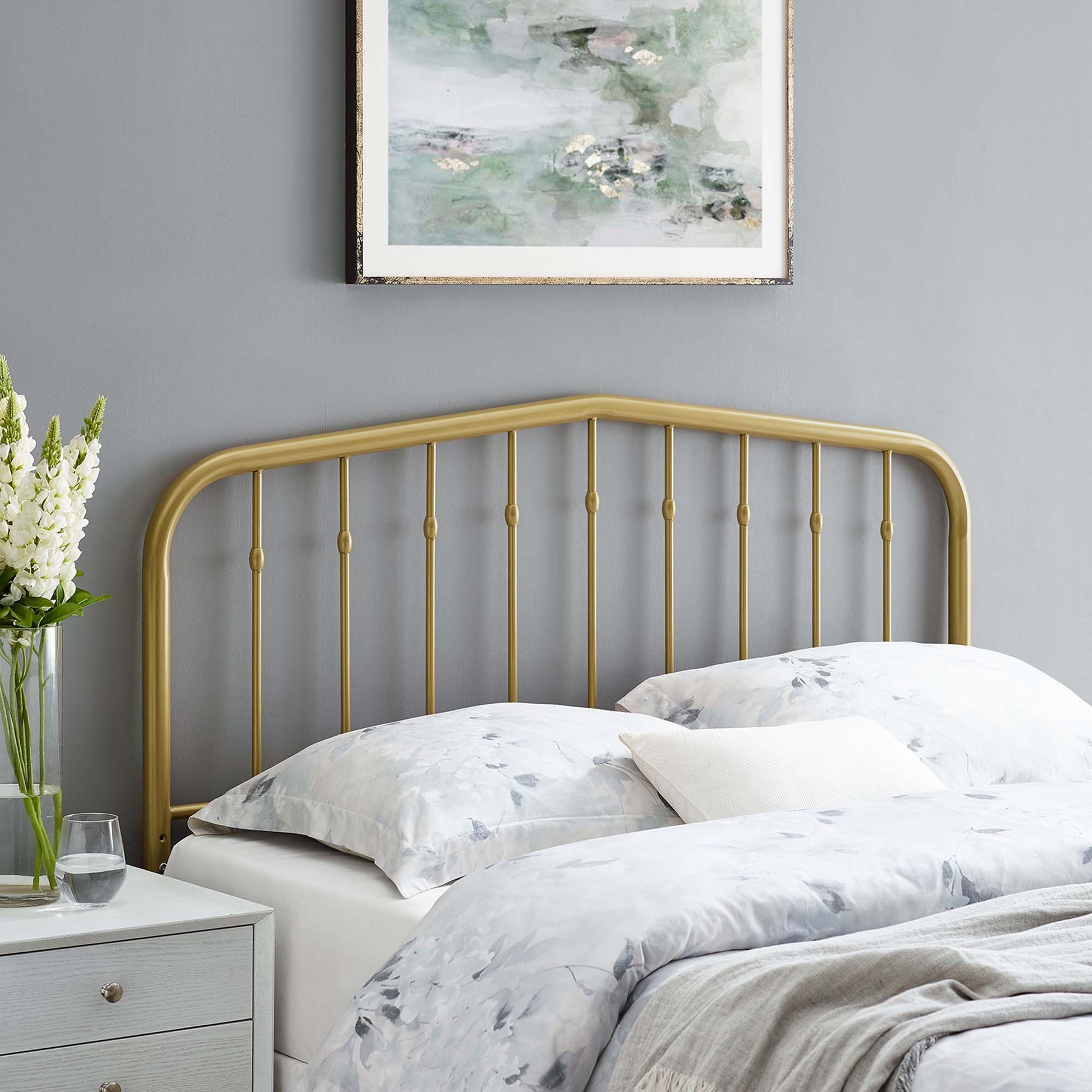 Yellow curved metal full headboard with vertical spindles, gray wall, abstract art, pale bedding, and a vase of flowers on a nightstand.