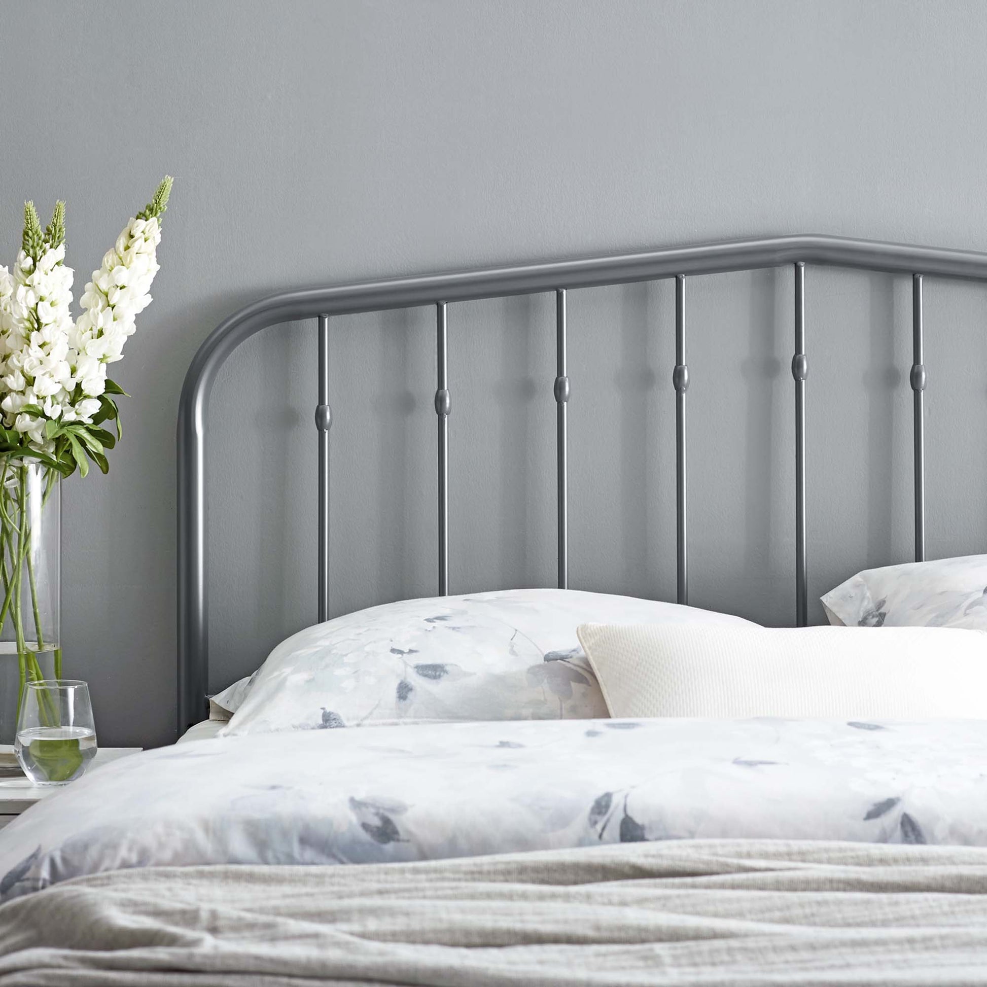 Grey metal headboard with a curved top rail and vertical spindles, placed on light bedding beside a vase of white flowers.