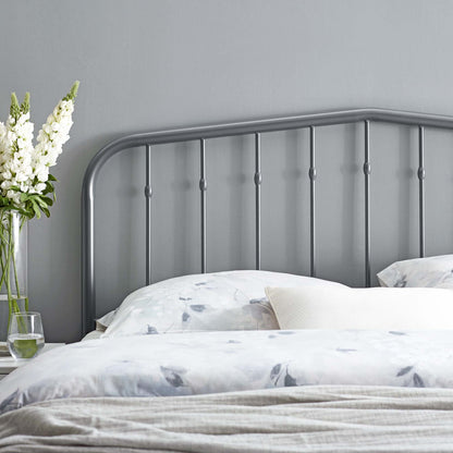 Grey metal headboard with a curved top rail and vertical spindles, placed on light bedding beside a vase of white flowers.