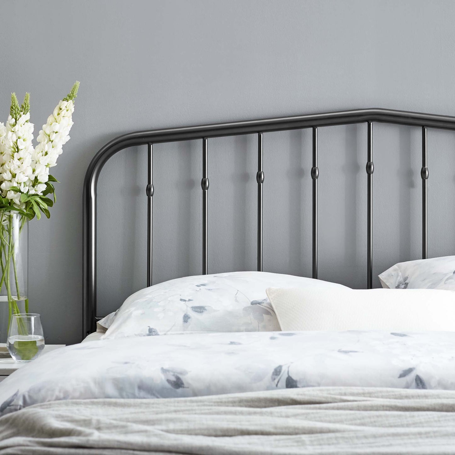 Black metal queen headboard with a curved top and vertical spindles, white floral bedding, gray wall, and a vase of white flowers.