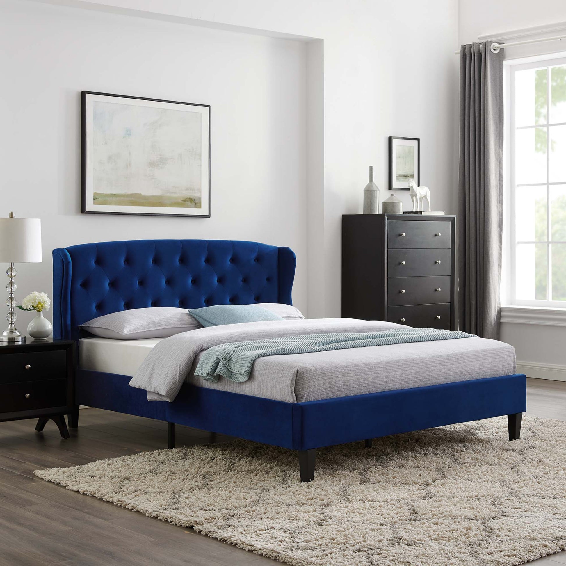 Navy tufted upholstered bed with headboard, gray bedding, black nightstands, dresser, window with curtains, cream shag rug.