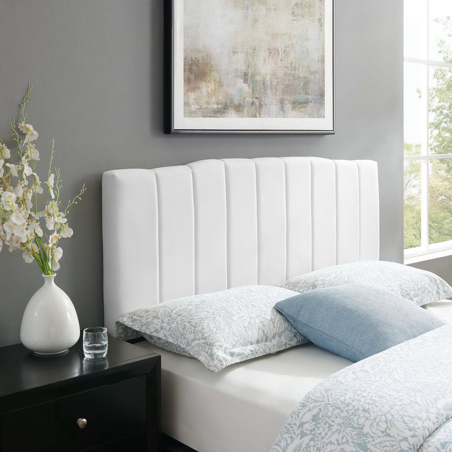 White upholstered headboard with vertical channels, pale bedding with blue pillows, dark nightstand with vase and glass.