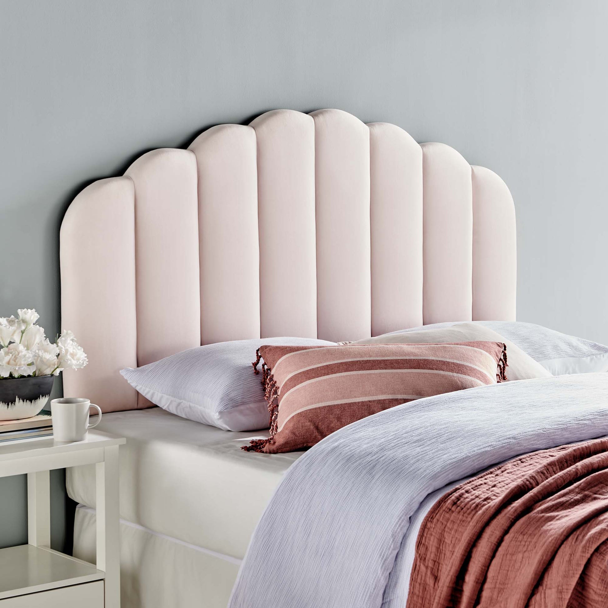 Red scalloped tufted twin headboard with white bedding, rust-red pillow and blanket, white bedside table with flowers.