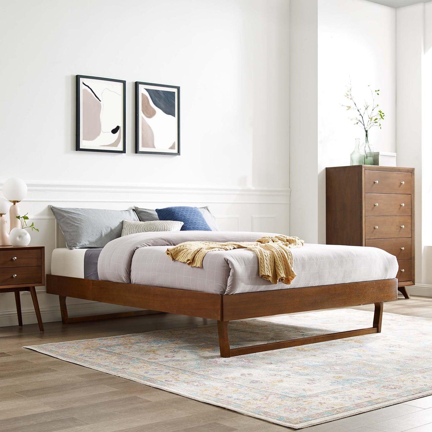 Walnut mid-century platform bed with gray bedding, blue pillows, mustard throw; nightstand, dresser, abstract art, pastel rug.
